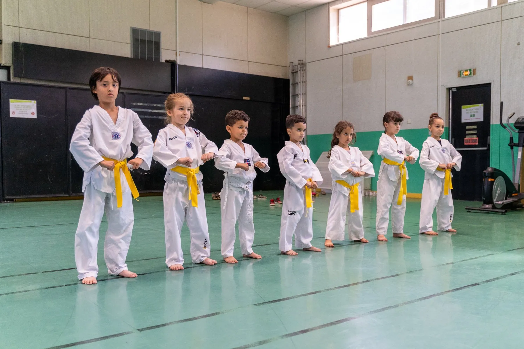Sept jeunes enfants en dobok, ceinture jaune ou blanche, alignés en position de garde dans le gymnase