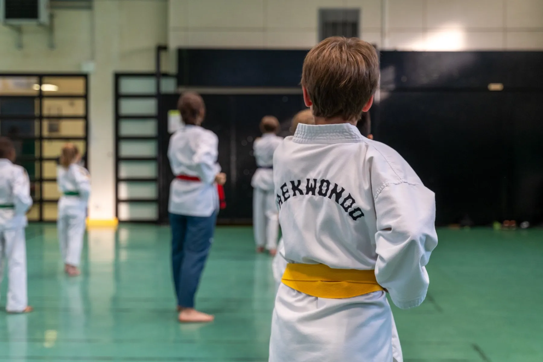 Un enfant de dos, « Taekwondo » brodé sur son dobok et ceinture jaune, suit le cours